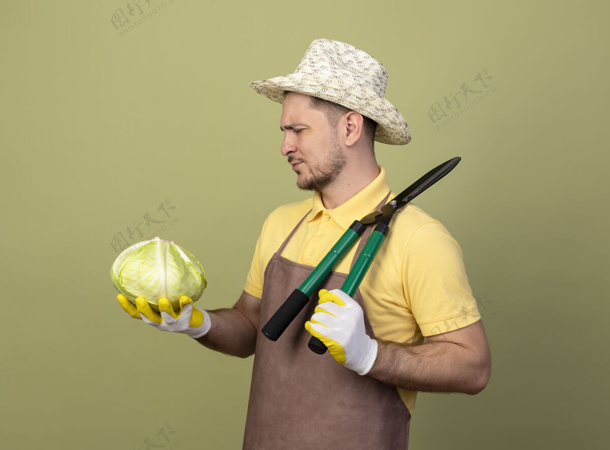 严肃一个年轻的园丁 穿着连体衣 戴着帽子 戴着工作手套 拿着树篱剪和卷心菜 严肃地看着它看男人连身衣