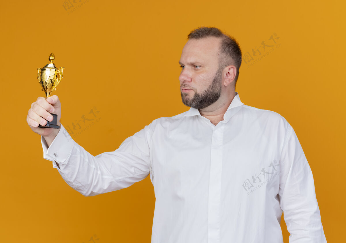 拿着留着胡子的男人穿着白衬衫拿着奖杯严肃地看着它站在橙色的墙上站着奖杯脸
