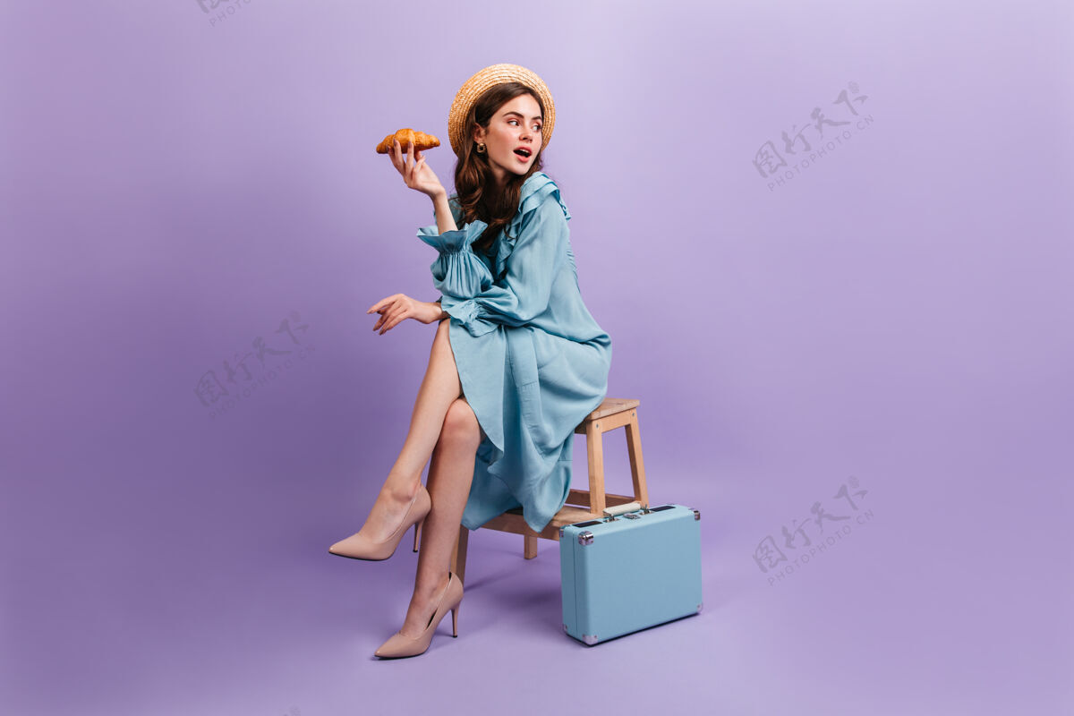 表情身着优雅蓝色连衣裙的年轻女士的全幅照片这位女士坐在手提箱旁边的凳子上 手里拿着美味的羊角面包有趣明亮女人