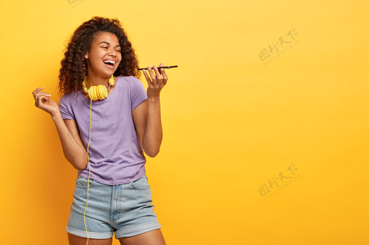室内摄影棚拍摄的快乐的年轻女性与非洲发型摆在黄色的墙壁通话欣喜若狂电话