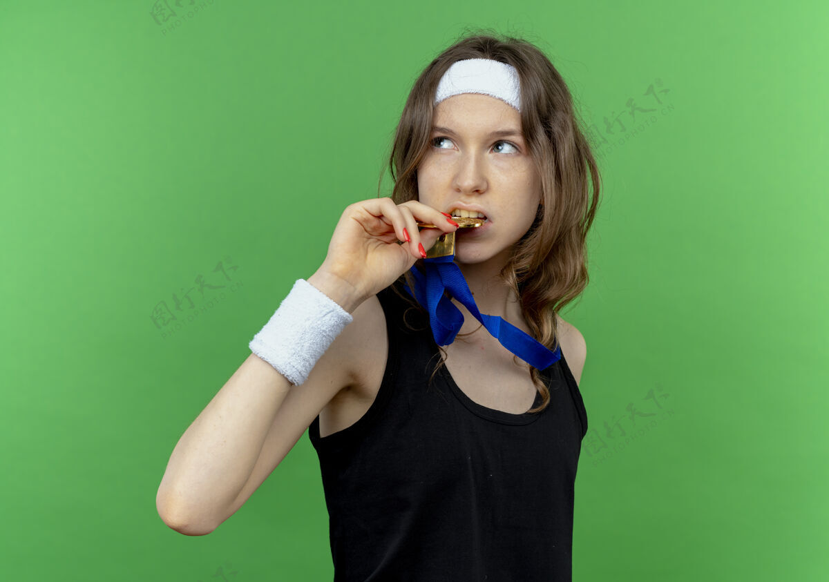 女孩身穿黑色运动服的年轻健身女孩戴着头带 脖子上挂着金牌 站在绿色的墙上一边看着一边咬着她的奖牌奖牌头带脖子