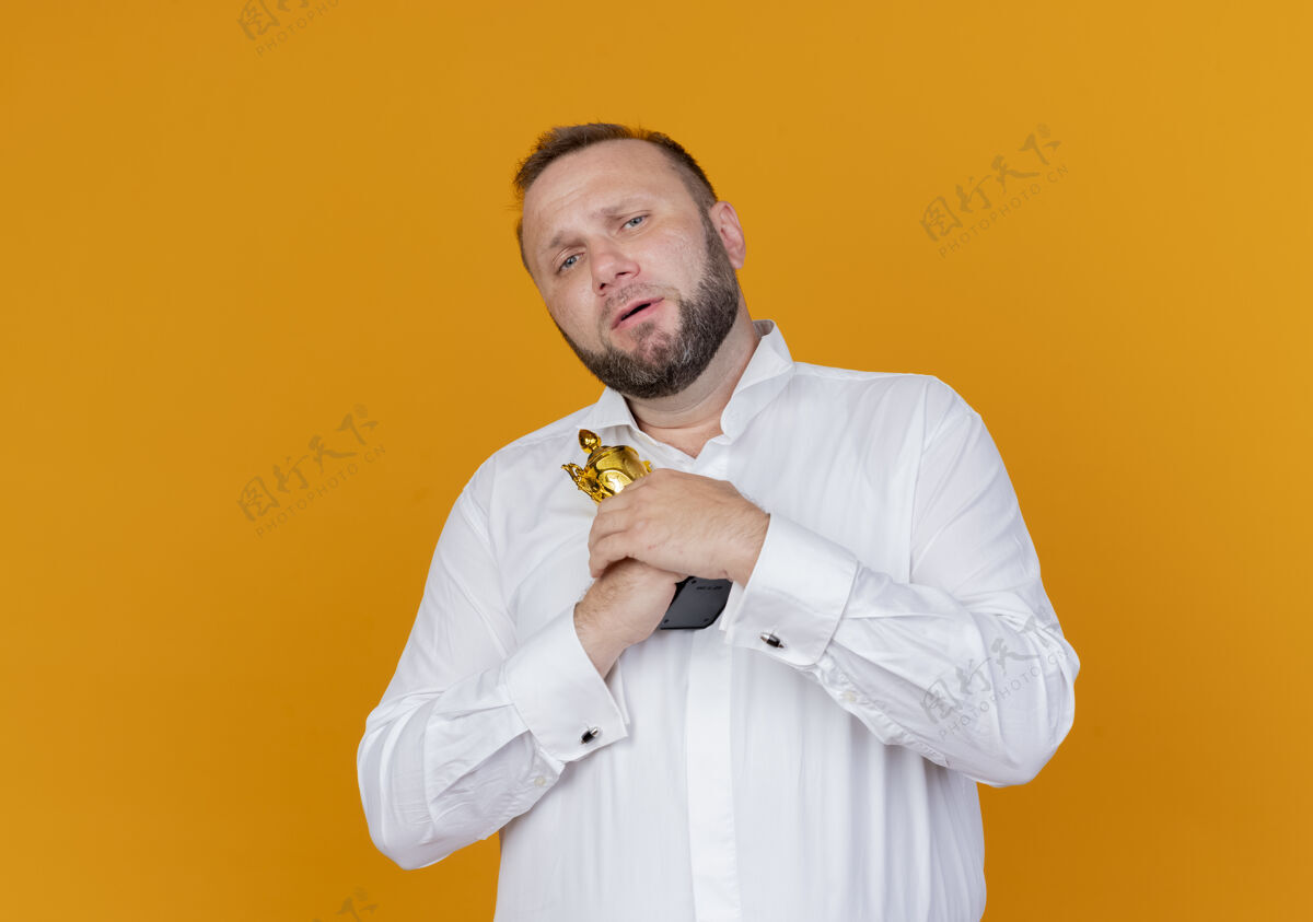 胡子身穿白衬衫 满脸胡须 手持奖杯的男子站在橙色的墙上 脸上露出不高兴的表情男人脸奖杯