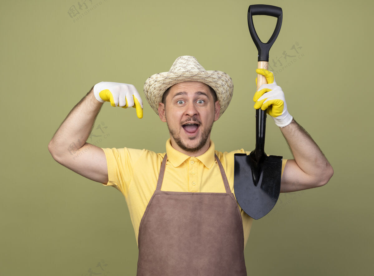 放下年轻的园丁 穿着连体衣 戴着帽子 戴着工作手套 拿着铲子指着地笑着手套连身衣穿