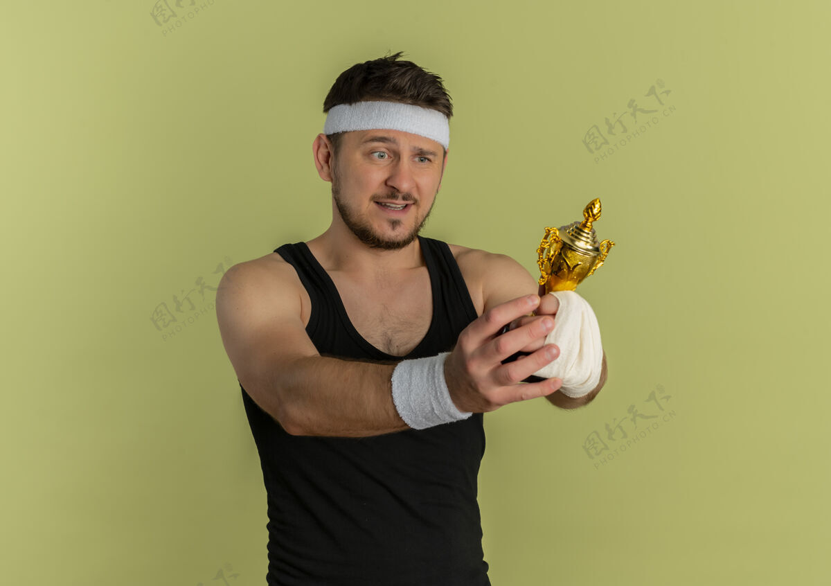 头带戴着头巾拿着奖杯的年轻健身男子站在橄榄色背景下兴奋而快乐地看着奖杯奖杯站兴奋