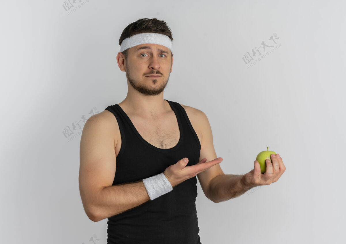 苹果戴着头巾拿着青苹果的年轻健身男子用胳膊哦手看着严肃的脸站在白色的背景上抱着脸站着