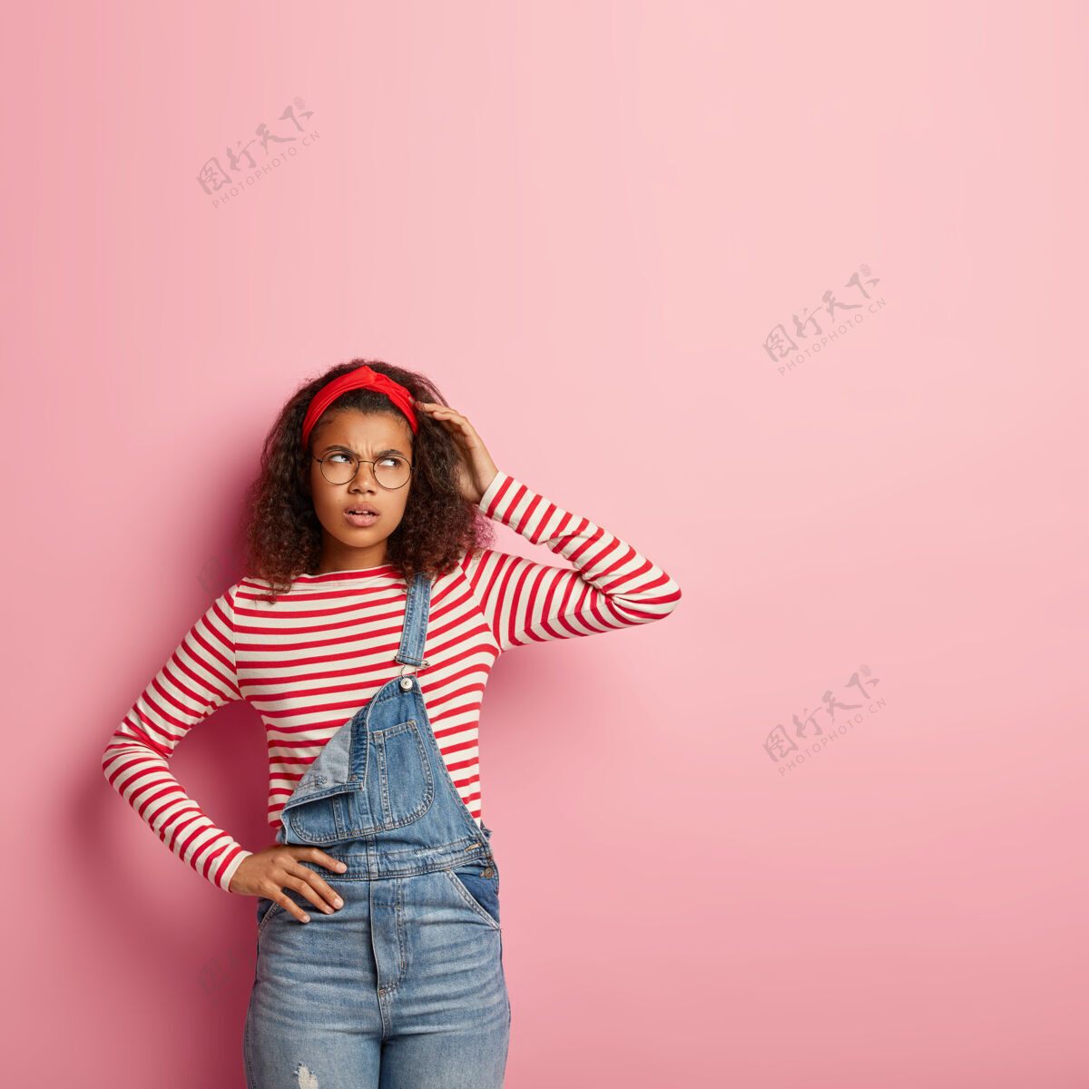垂直照片中 一位体贴 愤怒的非裔美国妇女 表情忧郁 搔着头 头上戴着红色头带 条纹套头衫和牛仔工作服思想女士年轻