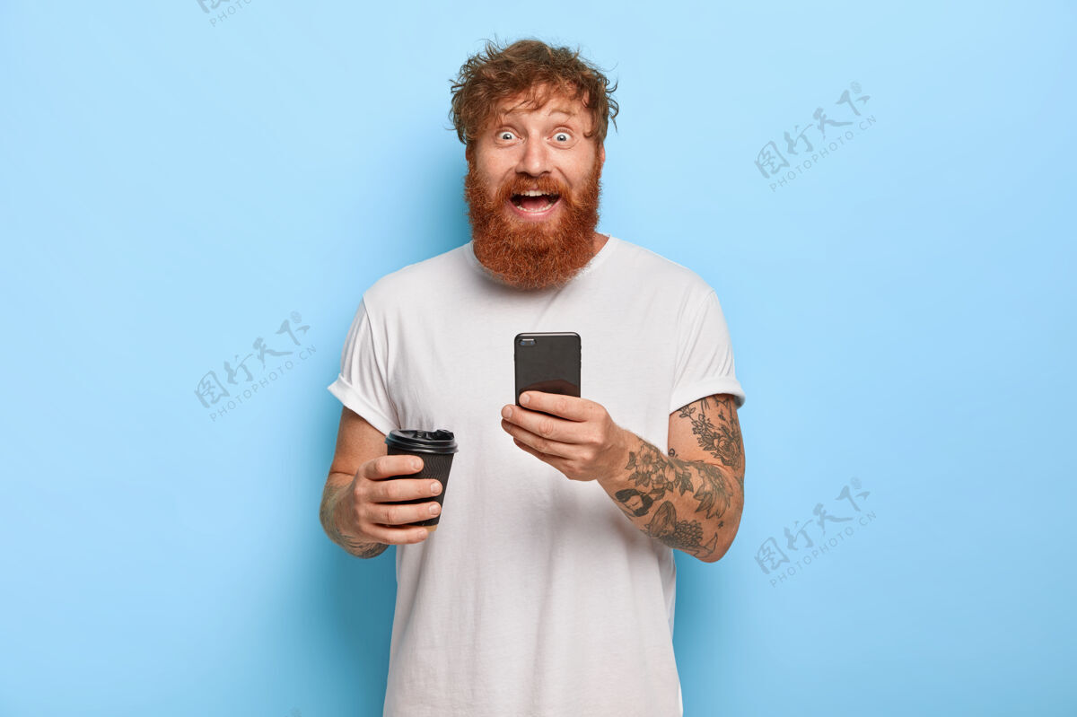 通知笑容可掬 满脸胡须 情绪激动的男人留着红发 拿着手机 和朋友分享好消息 面带笑容 眼睛瞪得大大的 穿着休闲的白色t恤 拿着外卖咖啡冲浪媒体服装