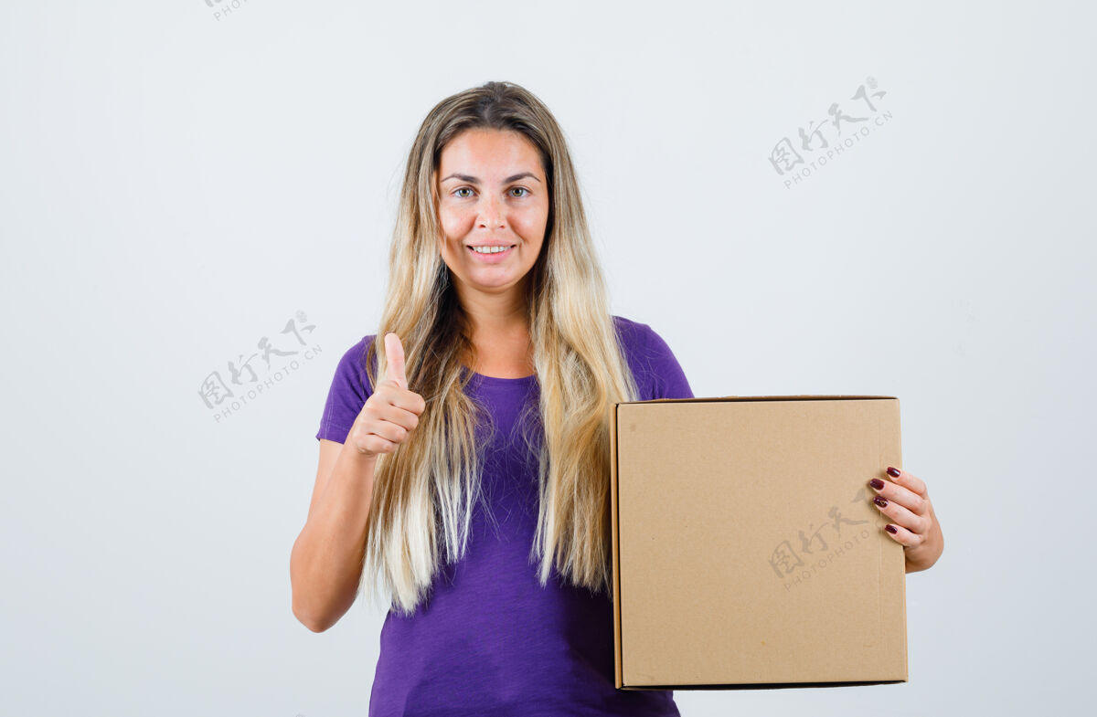 女人年轻的女性拿着盒子 穿着紫罗兰色的t恤 大拇指朝上 看起来很高兴 正面视图抱着高兴年轻