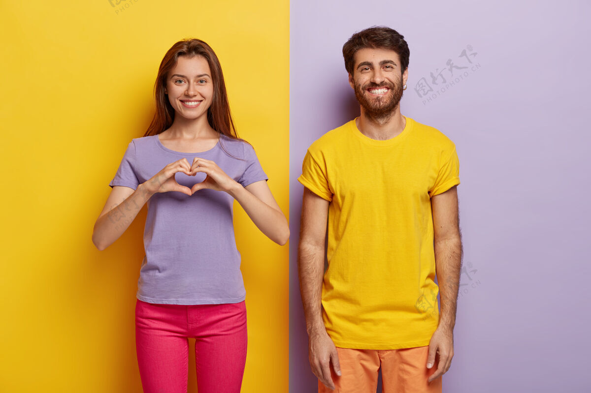 穿着积极的女人做心形 表达爱和美好的感情 她的男朋友站在旁边 带着牙齿般的微笑乐趣浪漫立场