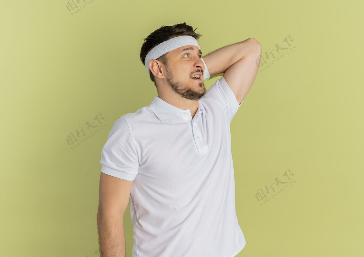 衬衫身穿白衬衫 头箍 困惑地站在橄榄色背景下的年轻健身男子白色看站