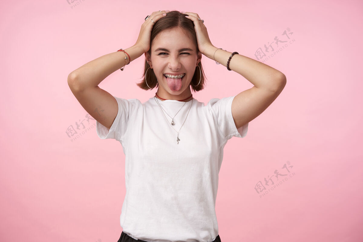 休闲摄影棚拍摄了一个快乐的年轻漂亮的短发黑发女子举着双手放在头上 愉快地伸出舌头 在粉红色的墙上摆姿势吊坠发型微笑