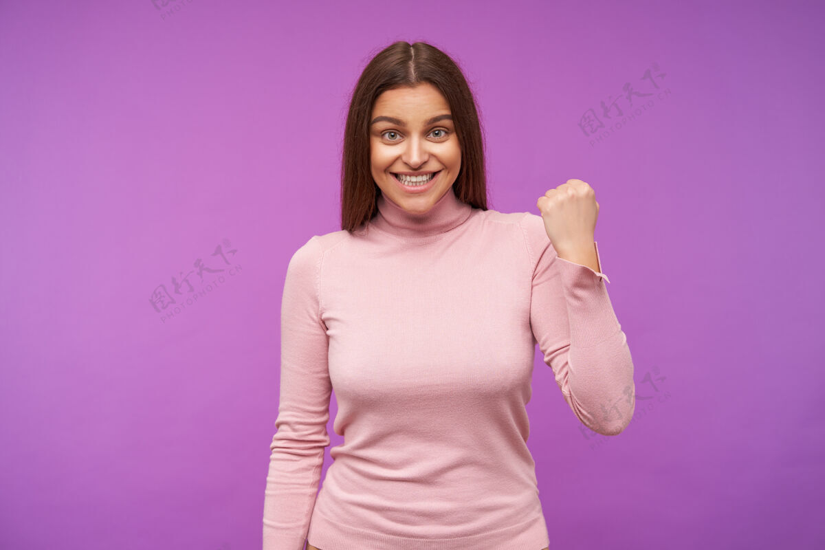 爱一个快乐的年轻碧眼黑发女人 她高高兴兴地举起她的拳头 面带微笑 穿着粉红色的马球衫 站在紫色的墙上心情漂亮黑发