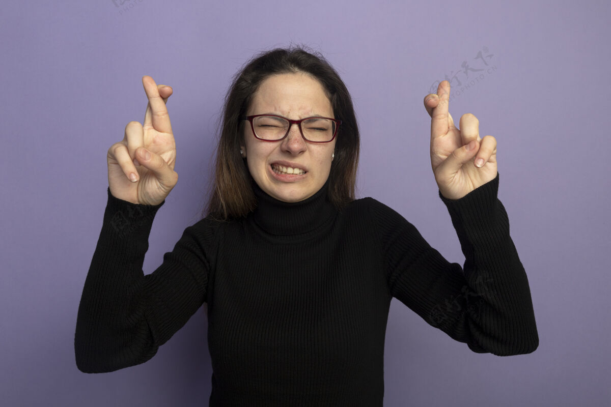 人身穿黑色高领毛衣戴着眼镜许下美好愿望的年轻美女闭着眼睛站在紫色的墙上 双手交叉着希望的表情年轻手指紫色