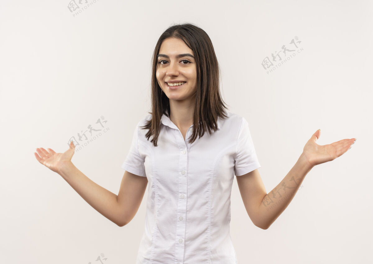 手势身穿白衬衫的年轻女孩望着前面 微笑着 愉快地向两边张开双臂 站在白色的墙上站欢呼人
