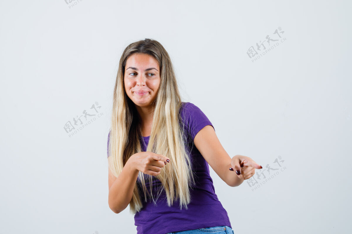 长金发女郎指着远处的紫罗兰色t恤 牛仔裤 看起来很高兴 前视图高兴人时尚
