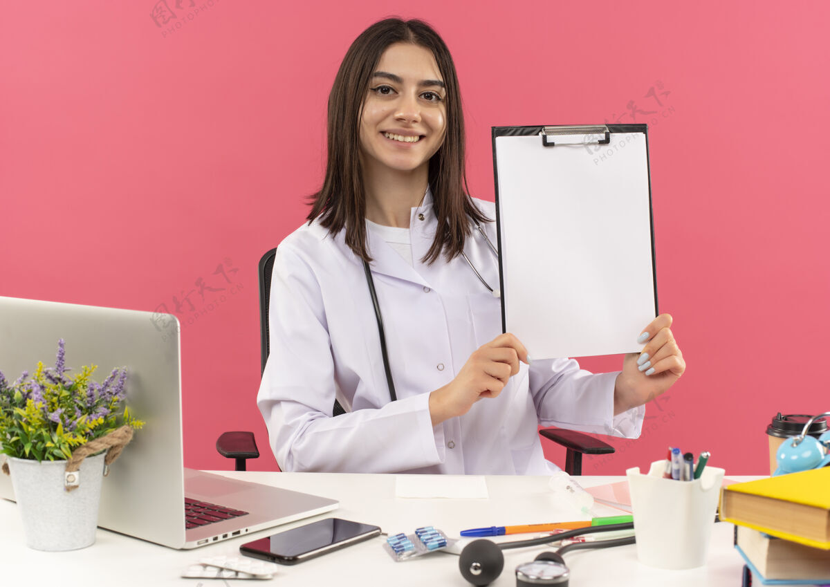 人年轻的女医生穿着白大褂 脖子上戴着听诊器 展示着空白页的剪贴板 微笑着看着前面坐在桌旁 笔记本电脑放在粉色的墙上桌子医院医疗保健