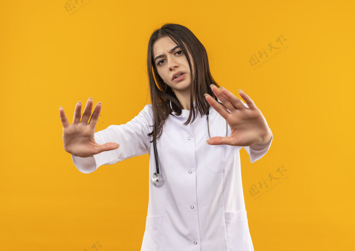 脸身穿白大褂 脖子上戴着听诊器的年轻女医生做着防御手势 双手朝前看 严肃的脸站在橙色的墙上年轻人外套医院