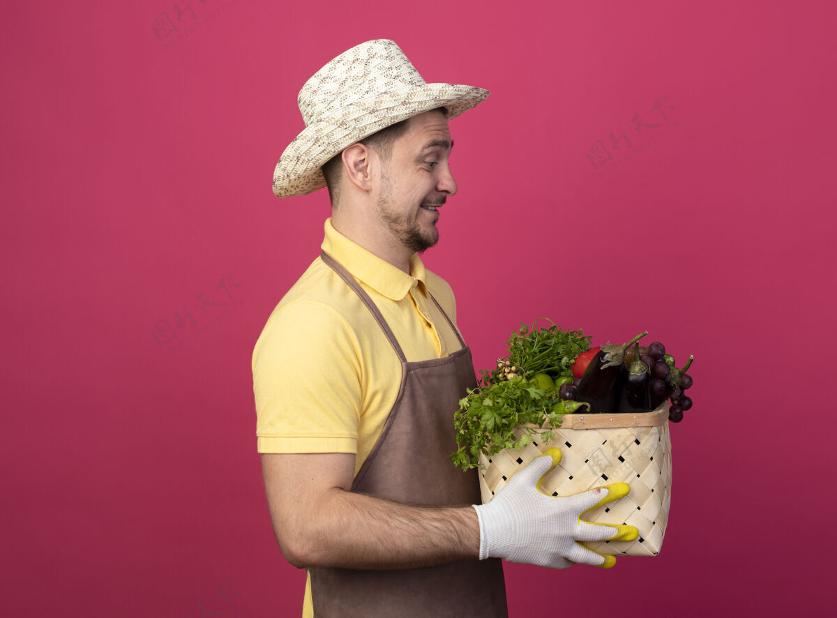 制服年轻的园丁穿着连体衣 戴着帽子 戴着工作手套 手里拿着装满蔬菜的板条箱 站在粉色的墙上 看着它们快乐而积极的样子粉色花园满的