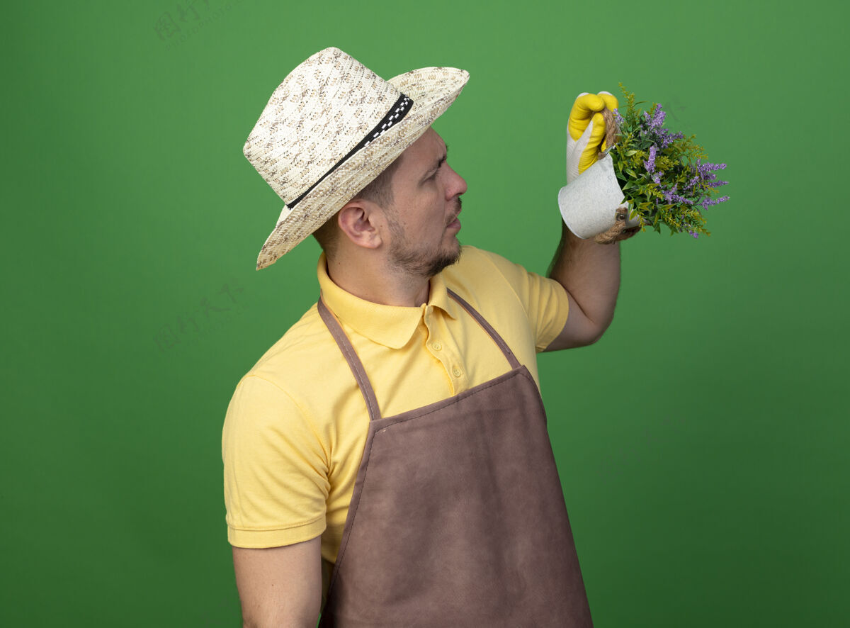 人年轻的园丁穿着连体衣 戴着帽子 戴着工作手套 手里拿着盆栽植物 站在绿色的墙上困惑地看着它制服人人