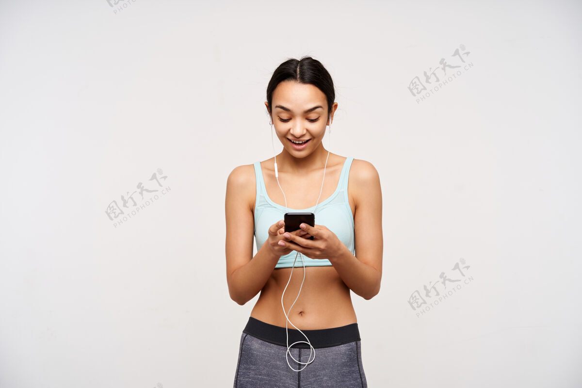 训练漂亮的年轻运动型棕色头发的女人穿着薄荷运动胸罩和灰色紧身裤手持手机 高兴地看着屏幕上 在白色的墙上摆姿势欧洲黑发锻炼
