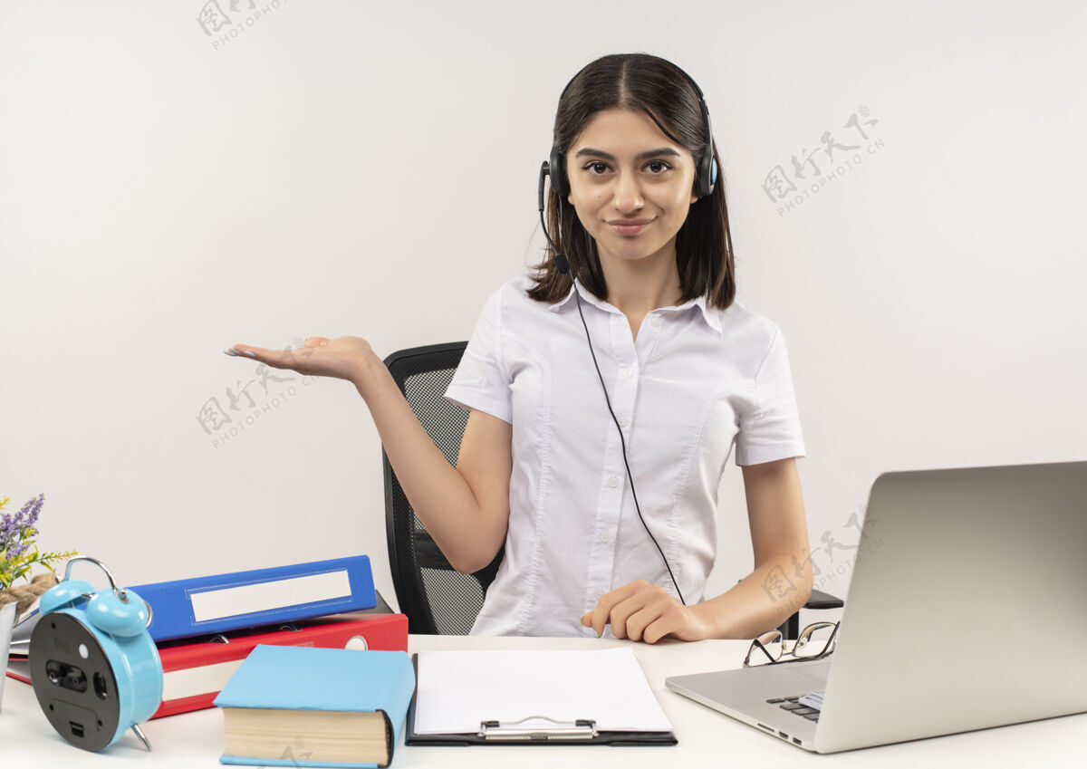 衬衫穿着白衬衫 戴着耳机的年轻女孩 手把手拿着东西微笑着坐在桌子旁 白墙上放着文件夹和笔记本电脑麦克风微笑桌子