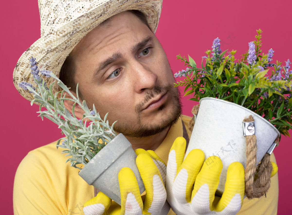 连身衣年轻的园丁穿着连体衣 戴着帽子 戴着工作手套 手里拿着盆栽植物 站在粉色的墙上看着它们困惑年轻手套困惑