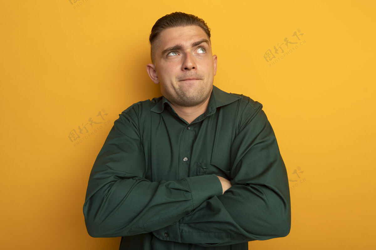 人身穿绿衬衫的年轻帅哥 双手交叉放在胸前 困惑地抬起头 站在橙色的墙上向上手臂橙色