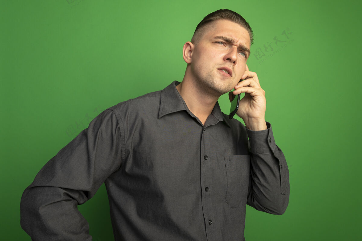 男人一个穿着灰色衬衫的年轻帅哥站在绿色的墙上严肃地讲着手机严肃年轻站立