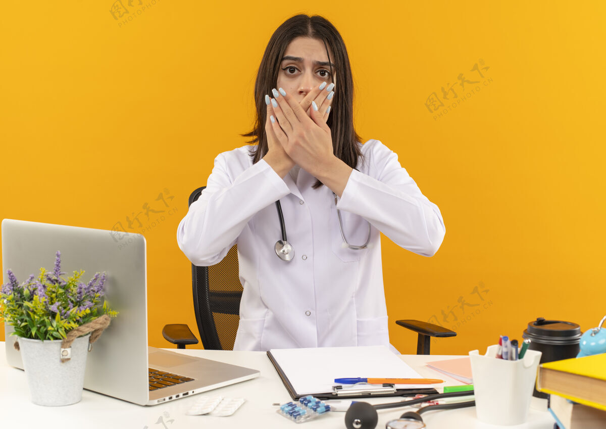 人身穿白大褂的年轻女医生戴着听诊器望向前方 双手捂着嘴巴坐在桌旁 手提电脑和文件盖在橙色的墙上手公民保健