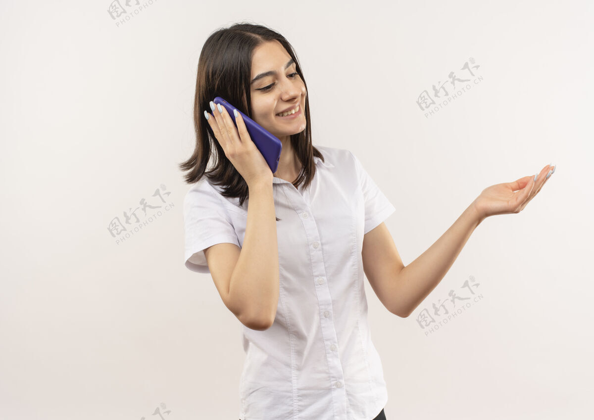 脸穿着白衬衫的年轻女孩站在白墙上 面带微笑地讲着手机女人站手势