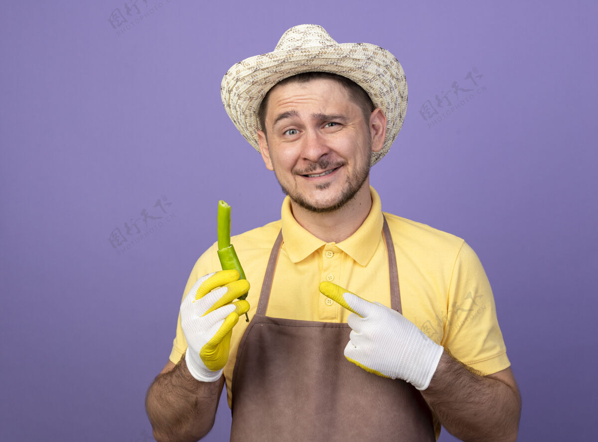 工作年轻的园丁穿着连体衣 戴着帽子 戴着工作手套 手里拿着破的青辣椒 用食指指着它 微笑着自信地站在紫色的墙上人制服穿