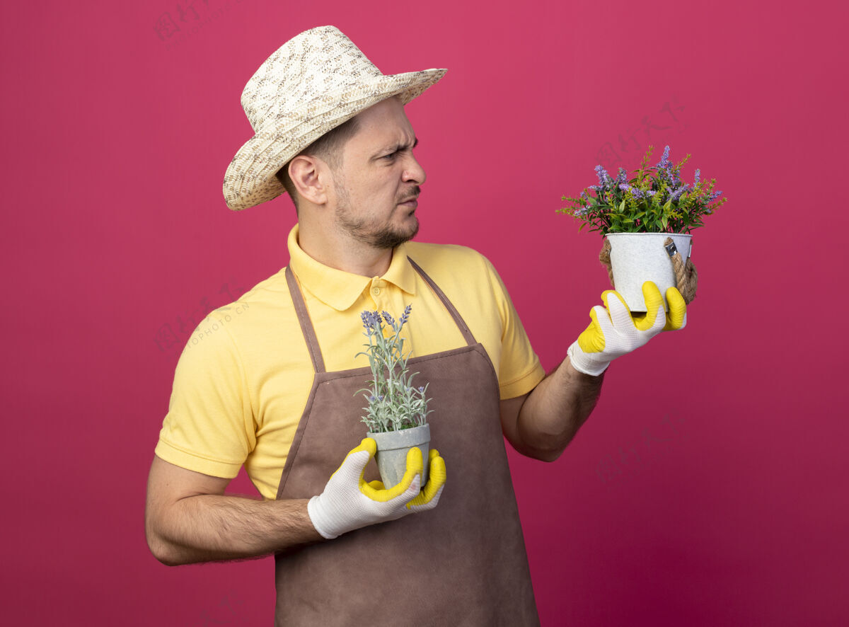 抱着年轻的园丁穿着连体衣 戴着帽子 戴着工作手套 手里拿着盆栽植物 看着他们站在粉色的墙上 困惑和不快人花园看