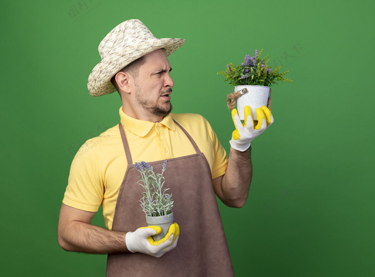 帽子年轻的园丁穿着连体衣 戴着帽子 戴着工作手套 手里拿着盆栽植物 看着他们站在绿色的墙上感到困惑和不快看人花园
