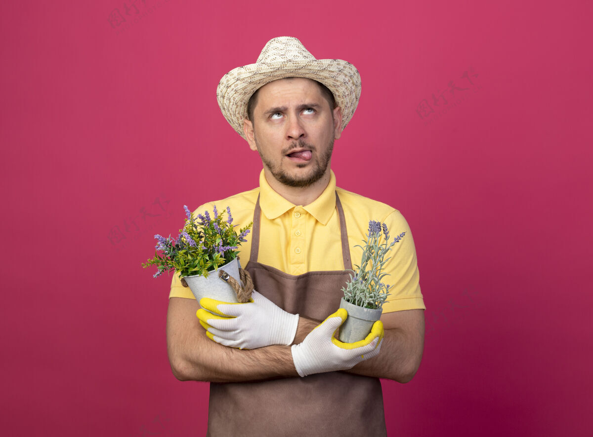 连身衣年轻的园丁穿着连体衣 戴着帽子 戴着工作手套 手里拿着盆栽植物 困惑地抬起头 伸出舌头站在粉红色的墙上人植物人