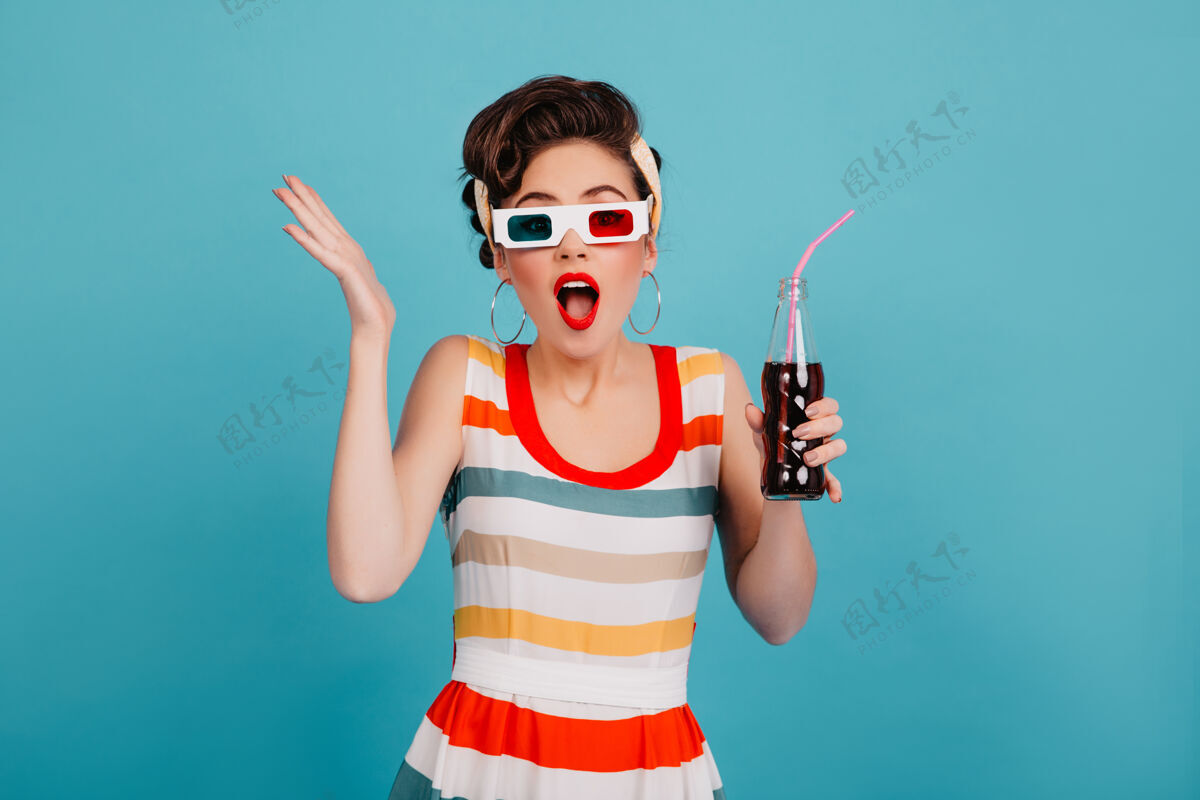 时尚一个戴着3d眼镜的震惊的年轻女子一个穿着条纹连衣裙的小女孩拿着一瓶苏打水 背景是蓝色的惊喜发型漂亮