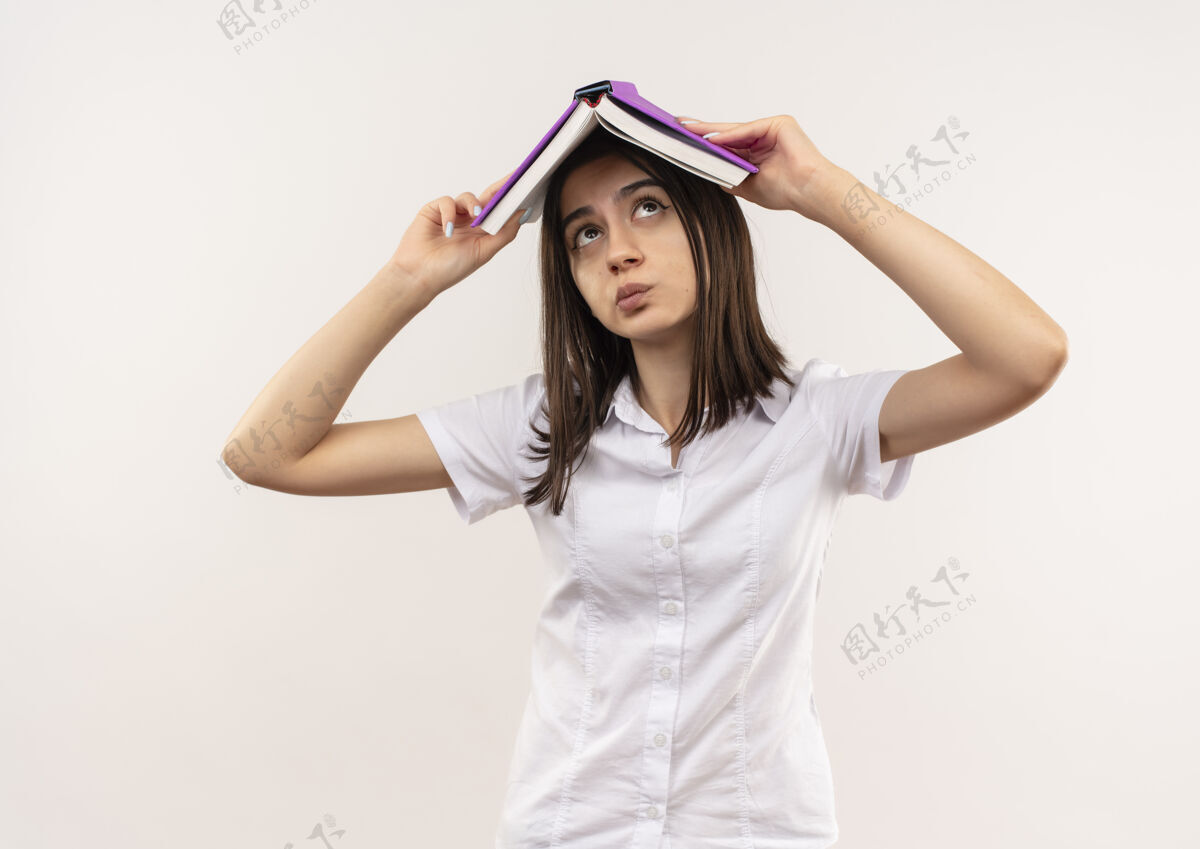 女孩穿着白衬衫的年轻女孩头上举着一本打开的书 站在白色的墙上 看上去很困惑女看开