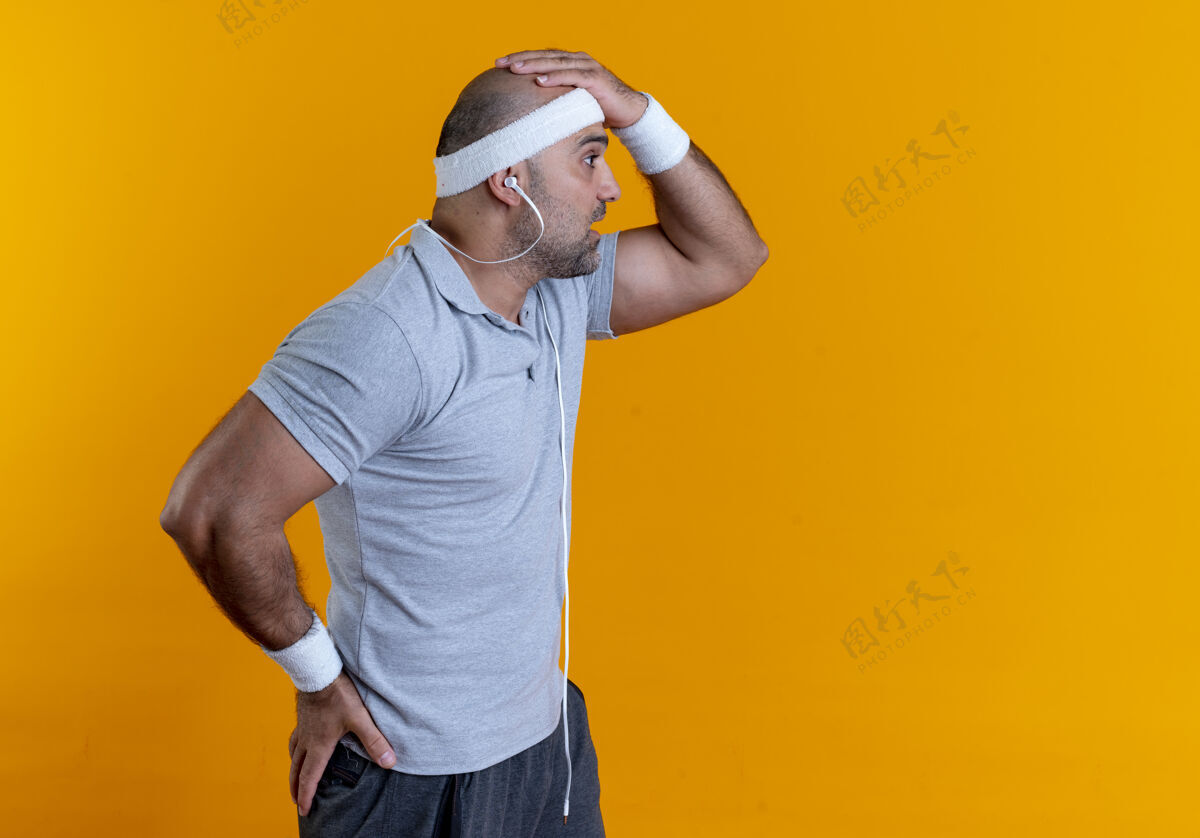 姿势戴着头巾的成熟的运动型男人站在橙色的墙上 一边看一边 表情困惑肌肉男人人