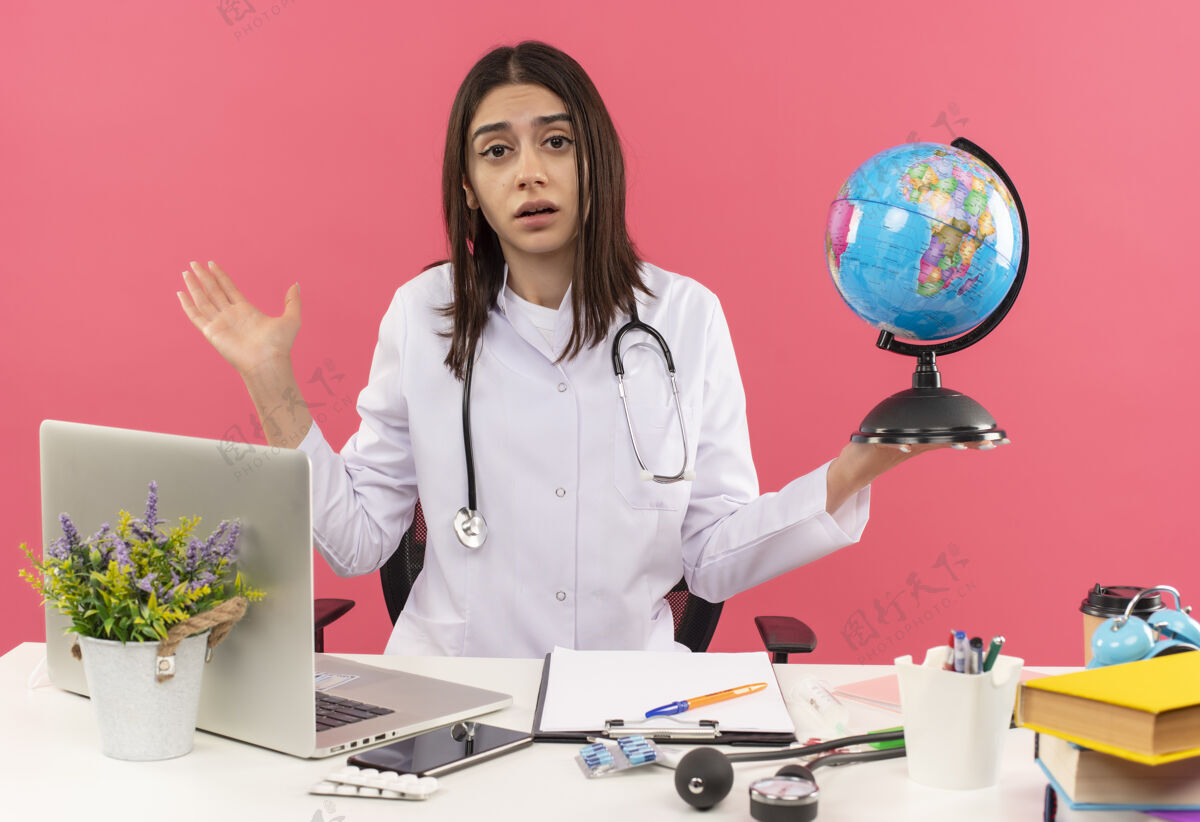 周围年轻的女医生穿着白大褂 脖子上戴着听诊器 手里拿着地球仪 看起来很困惑 耸耸肩 坐在桌旁 粉色墙上挂着笔记本电脑工作看医院