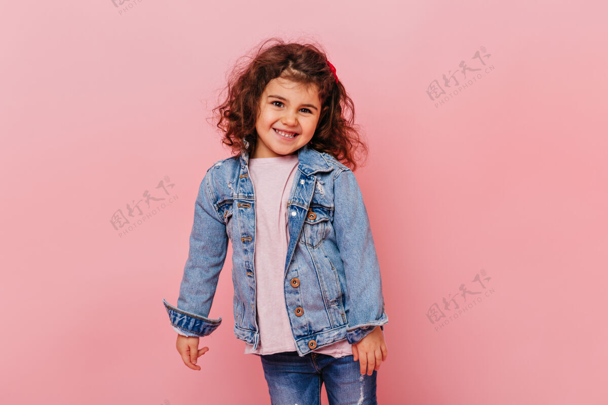 孩子微笑的小女孩 波浪形的头发 站在粉红色的背景上摄影棚拍摄的可爱的未成年儿童穿着牛仔夹克牛仔夹克肖像可爱