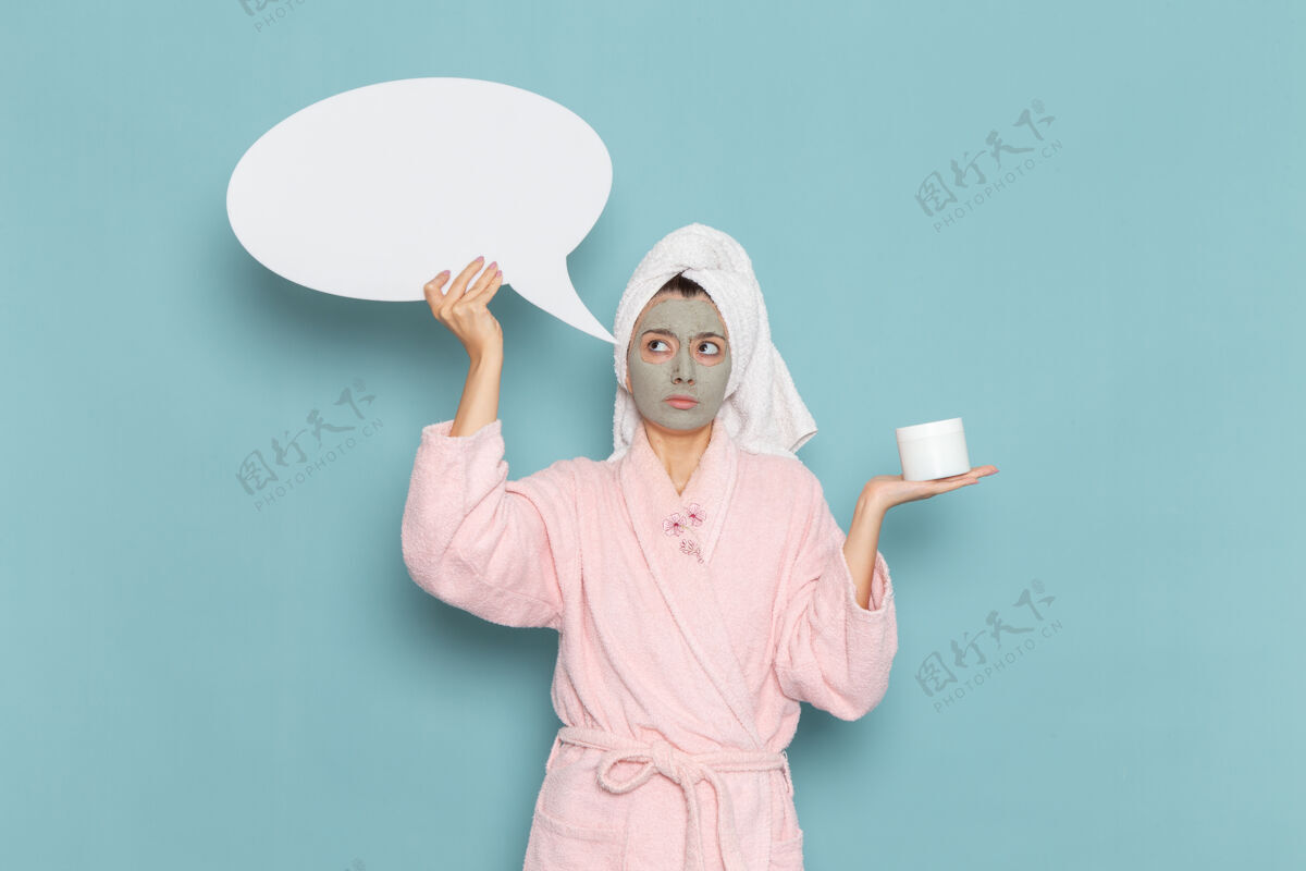 浴袍正面图年轻女性穿着粉色浴袍淋浴后手持巨大的白色标志在蓝色墙壁上美容水霜自理淋浴浴室成人人自理