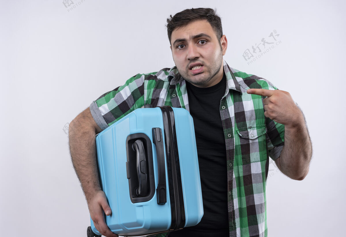 衬衫穿着格子衬衫的年轻旅人拿着手提箱 困惑地用手指着站在白墙上的自己手提箱看困惑