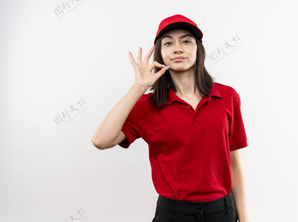 微笑年轻的送货员身穿红色制服 戴着帽子 带着怀疑的微笑 表现出沉默的姿态 就像在白色的墙上拉着拉链闭上了嘴穿女孩帽子