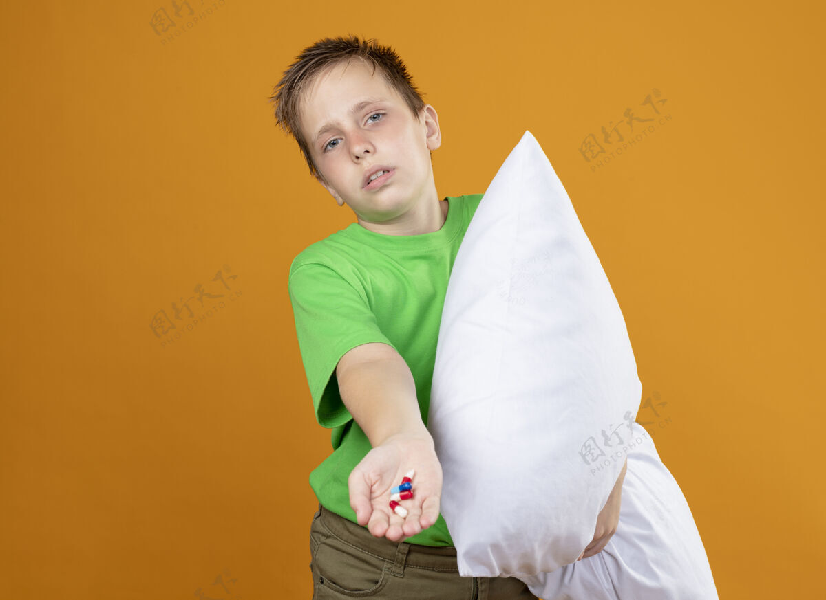 枕头生病的小男孩 穿着绿色t恤 感觉不舒服 抱着枕头 手里拿着药片 站在橘色的墙上 看起来很困惑 不开心 生病生病不舒服生病