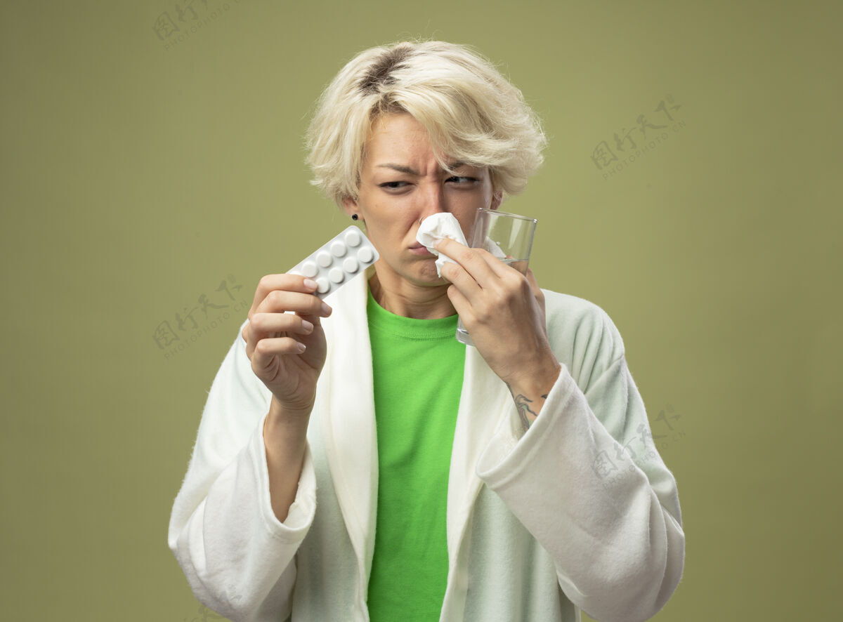 女人一个短发的生病的女人拿着一杯水和一个水泡 拿着药片 站在光墙上用餐巾擦鼻子光不适鼻子