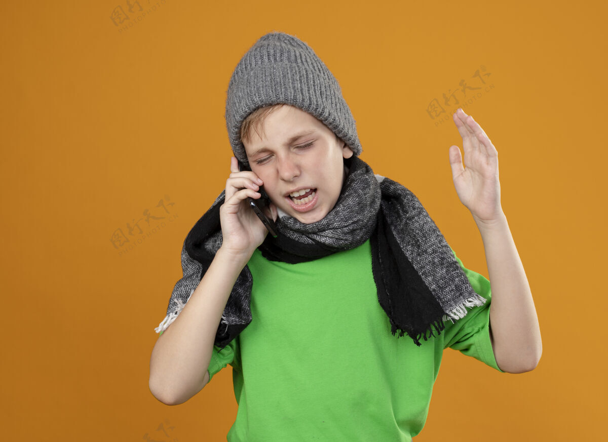 疾病生病的小男孩穿着绿色t恤 戴着暖和的围巾 戴着帽子 不舒服地讲着手机 站在橘色的墙上不高兴的表情烦恼温暖橙色