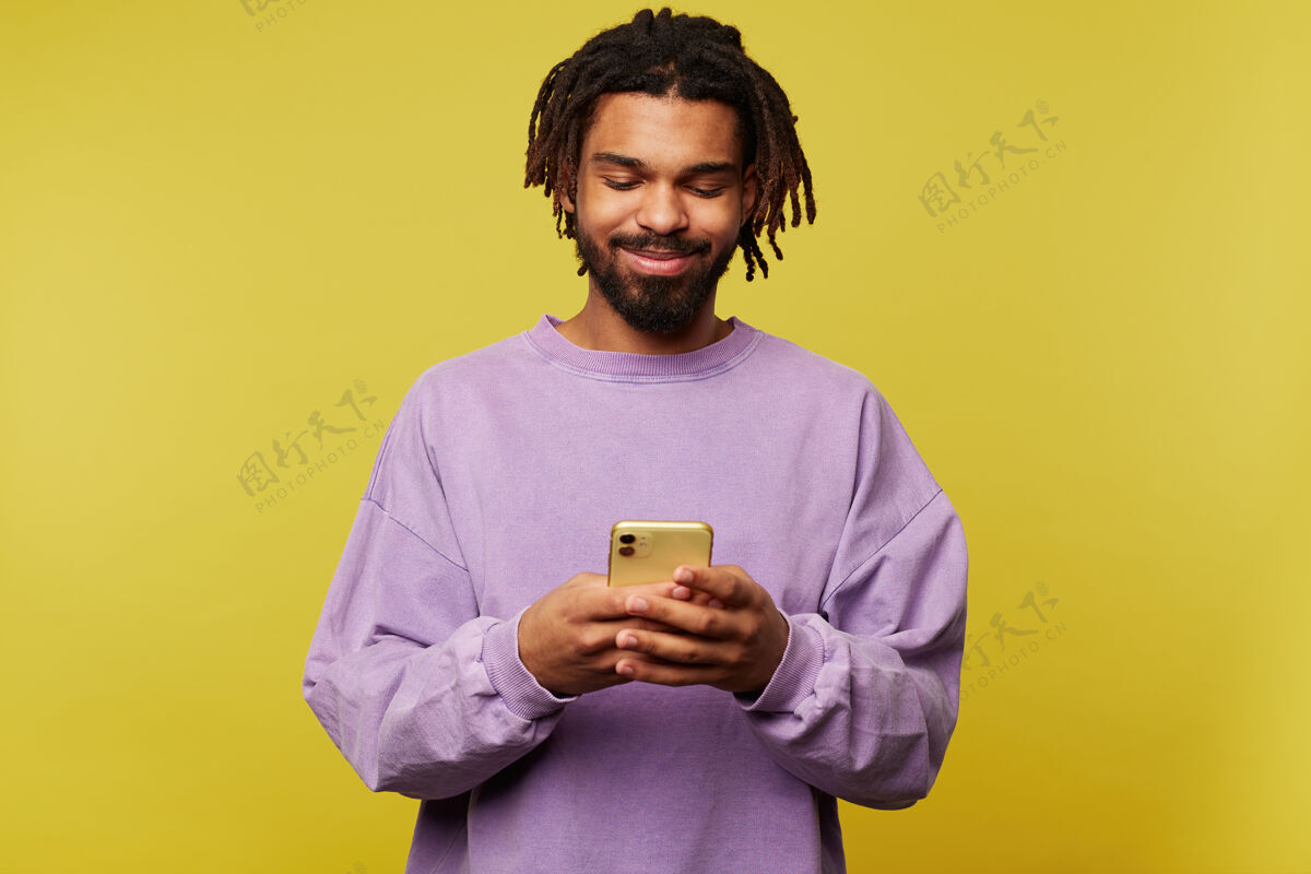穿着一张室内照片 年轻的黑皮肤黑发男性高举着智能手机 积极地看着屏幕 隔离在黄色背景下理发运动衫站立