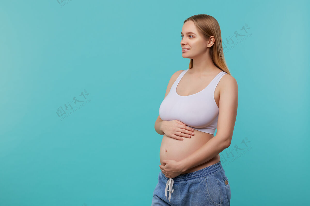 白头一张室内照片 年轻迷人的白头孕妇举起双手放在肚子上 面带微笑沉思地看着一边 在蓝色背景上摆姿势上衣金发表情