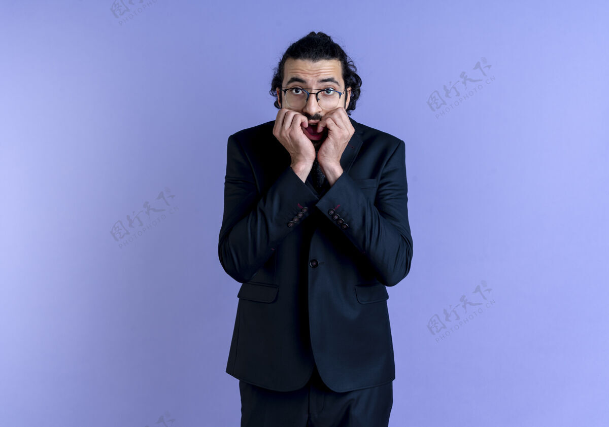 紧张一个穿着黑色西装戴着眼镜的商人站在蓝色的墙上 看着前面紧张而紧张的钉子西装姿势站