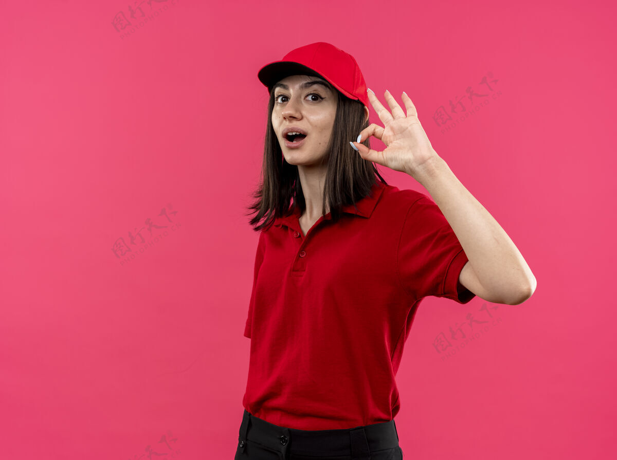 马球穿着红色马球衫 戴着帽子 微笑着 粉色墙上挂着“ok”的牌子 年轻的送货员很高兴显示交货帽子