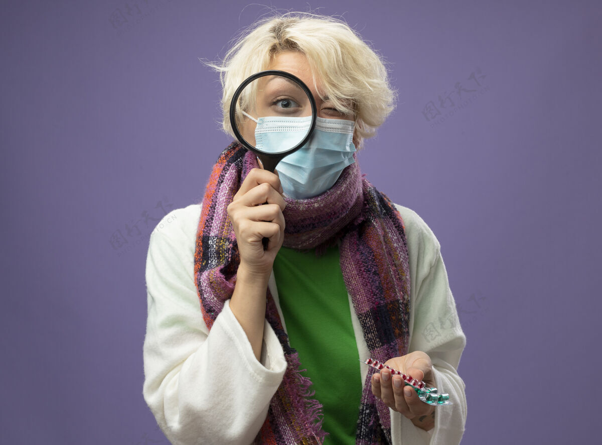 持有生病的不健康的女人 短头发 戴着暖和的围巾 戴着面罩 通过放大镜拿着药丸站在紫色的墙上玻璃通过头发